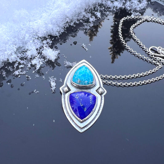 Hand crafted necklace with a pendant featuring turquoise and lapis stones on a reflective surface with snow.